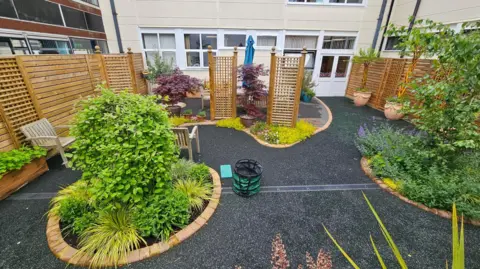 MCHT A garden behind a white hospital building. The area is mostly tarmac and is bordered with a brown, wooden fence. There are planted areas dotted around, including two large bushes in the middle, with smaller plants around the border.