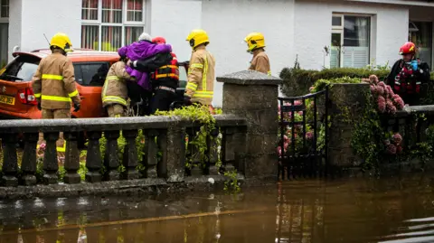 Pacemaker An elderly person is being lifted by fire service into a car as water flows around her home.