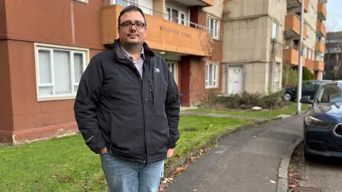 Dimitros standing in front of the entrance to Westfield Court. He has dark hair and dark-framed glasses. He is wearing a dark jacket over a light shirt and jeans.