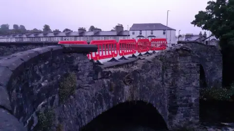 A row of five plastic fence segments have been erected along Nether Bridge. The wall of the stone bridge has been destroyed. A road of houses can be seen in the background.
