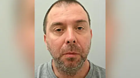 Police custody image of David Shorrock with brown thinning hair and brown and grey beard wearing a grey top.