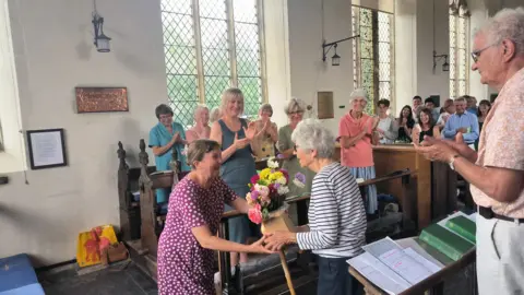 Cressi Sowerbutts An elderly woman is gifted flowers by another woman in the aisle of a church. People standing in the pews are watching on, smiling and clapping. 