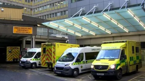 Four parked ambulances sit outside the main doors of the emergency department at the Royal Victoria Hospital in Belfast. Two of the vehicles are yellow and two are green. Two futher vehicles can be seen in the background, parked under a roof.