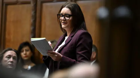 House of Commons Leigh Ingham - a woman with glasses, long dark hair and wearing a purple jacket over a white top - stands and speaks in the House of Commons while reading from a piece of paper.
