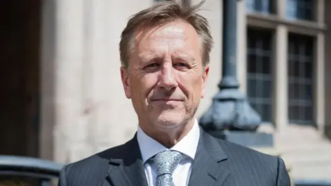 A head and shoulders image of Martin Murray in a grey suit and silver coloured tie standing in front of a council building