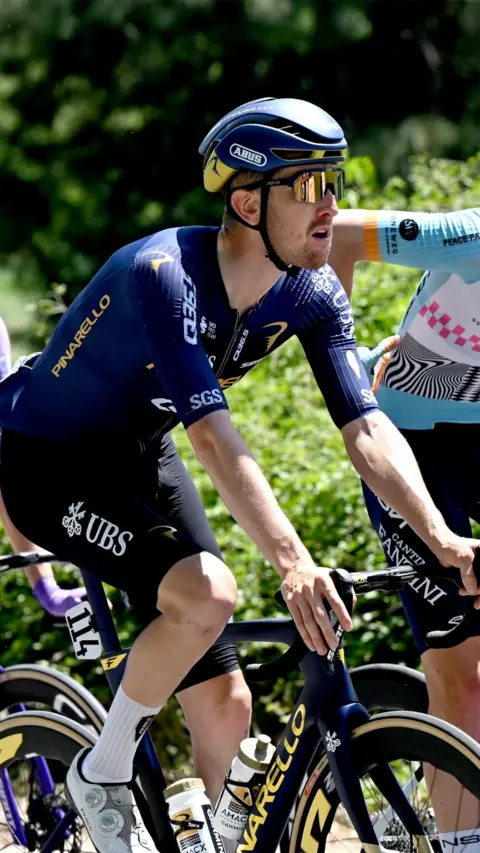 Close-up of a cyclist riding a road bike during a race, wearing a dark blue team kit, helmet, and mirrored sunglasses, with another rider partially visible alongside and green bushes blurred in the background.