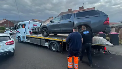 South Yorkshire Police A large wagon with a trailer on the back which has a grey pick-up truck on it. There are two men stood by the side, one is wearing black trousers and a top with 'Police' written on the back in white writing. 