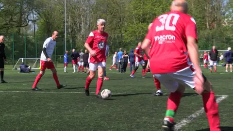 Men in red and white walking on an Astro turfed football pitch with a football at one man's feet. They are playing a match.