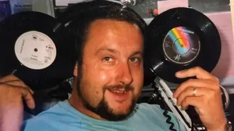 John Randall John Randall sitting in a radio studio wearing headphones and holding up two records.