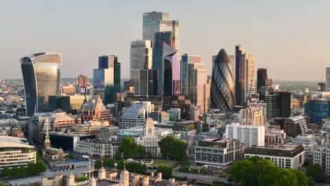 View of the major buildings of the City of London, headquarters to many financial firms, at dawn. The low sun off screen to the right illuminates the facades of the many multistorey skyscrapers, with the turrets of the Tower of London in the foreground.