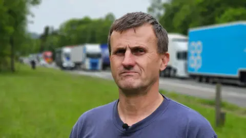 A man with short brown hair and a blue tshirt stands at the side of a road with stationary lorries behind him