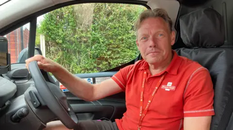 A man is sitting in the driver's seat of a vehicle. He is wearing a red polo shirt with the Royal Mail logo on the left side of his chest.