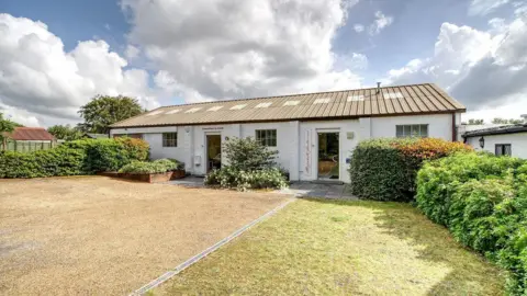 A single storey white building with a sloped roof. It is pictured on a clear day and has green grass and manicured hedges in front of it.