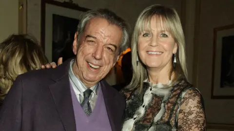 PA Media A couple smile as the stand in a dimly lit room. The man on the left has grey hair, swept back, and wears a dark blazer over violet sweater, striped shirt and grey tie. The woman has long blonde hair with a fringe and wears a lacy top.