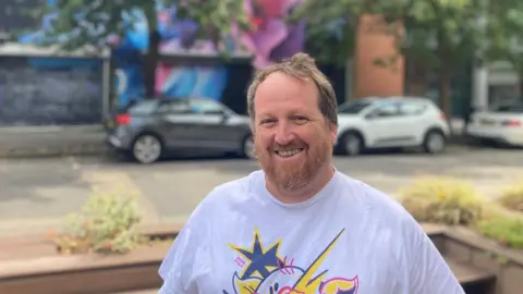 A man in a white t shirt smiling at the camera. He has a brown beard and brown hair. Behind him are some parked cars. 