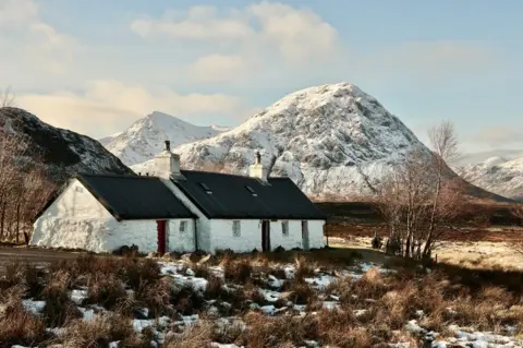 Graham Christie A small white cottage with a dark roof sits on frosty ground in front of a snow covered mountain peak