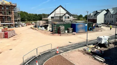 A house in the middle of a sandy building site, covered in scaffolding. In front of it are green and blue containers. On the right, some completed properties, and on the left is a property being built. 