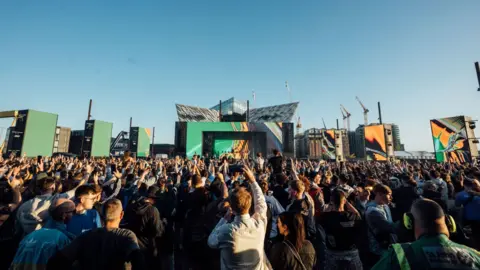 The Hype Factory A crowd of people standing recording and watching an artist play on stage at a festival. The image is taken from behind the crowd.
