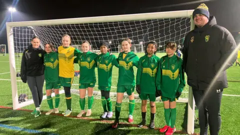 Elizabeth Baines / BBC A girls football team stands in front of a net. They wear green suits.