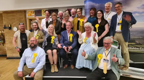 A group of Liberal Democrat Councillors smiling and waving at the camera in celebration. Some of them are sitting on a stage, some are sat on chairs and others are standing in the background. They are all wearing yellow rosettes on their chest.
