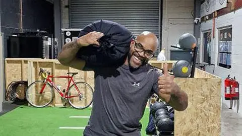 Daniel Jude Daniel Jude wearing a grey t-shirt carrying a 50kg sandbag on his shoulder holding his thumb up smiling in a gym