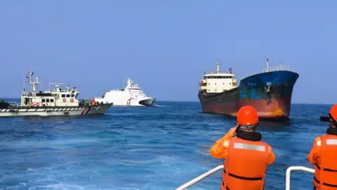 Back view of men in bright orange vests and helmet standing at the tip of the ship. In front of them are three more ships.