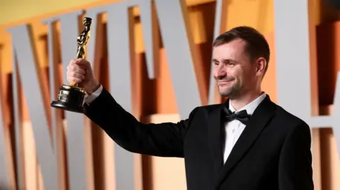 Getty Images Rhys Salcombe attends the 2025 Vanity Fair Oscar Party wearing black tuxedo and holding up Oscar statuette smiling