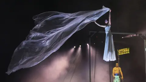 Yui Mok/PA Media A woman wearing a long white dress standing on a tall stilt on stage. She is holding up a large floaty piece of white material.