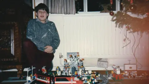 Simon Papouis A childhood photo of Simon Papouis wearing a blue jumper crouched next to a Lego train set. He is to the left of a Christmas tree, and is in front of a white radiator.