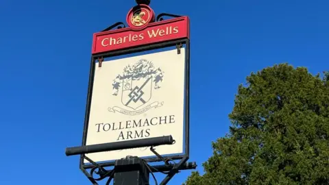 Steve Hubbard/BBC Pub sign on top of a post for the Tollemache Arms showing a coat of arms on a white background, and red Charles Wells section above the swinging bit of the sign.