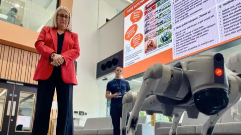 Ant Saddington/BBC A woman wearing glasses and a red jacket and dark blue top and trousers looking down at a four legged robot being operated by a student in the background. 