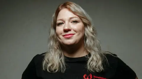 Underground standup Hana Kochehura, a blonde wearing winged eyeliner, smiles at the camera while wearing a black T-shirt. 