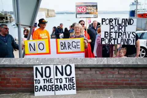 PA Media Protestors stand in front of Portland docks with placards with slogans including 'No to the barge' and 'Portland Port betrays Portland'