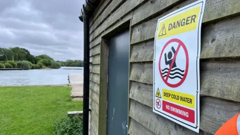 A sign warning people of deep cold water and telling them not to swim is nailed to a wooden shed beside a lake.