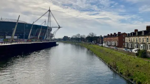 A stretch of the river Taff which passes the Principality Stadium to the left and terraced houses along the embankment to the right. 