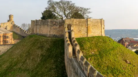 Universal Images Group/Getty Images A low stone castle tower with a large circumference atop a green mound, with battlements to either side. Within the tower, which is roofless, trees are growing. Houses can be seen beneath the mound to the right.