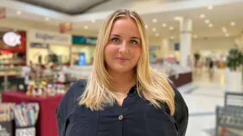 BBC Zoe Bulter smiles looking directly at the camera, dressed in a black shirt with long blonde hair standing in a shopping centre