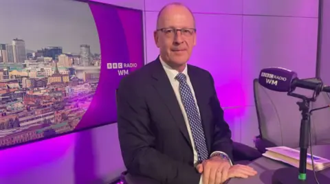 Person wearing a dark suit, white shirt, and blue polka-dot tie seated in a radio studio. A BBC Radio WM microphone and chair are visible beside a desk, with purple studio lighting and a cityscape graphic featuring the BBC Radio WM logo displayed on a screen in the background.