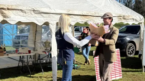 A man on the right wearing a cap and jacket is holding the bouquet along with a woman with blonde hair wearing a blue top and jeans.