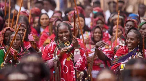 As mulheres Amensisa IFA / BBC carregam palitos de madeira esculpidos e marcam em roupas vermelhas combinando.