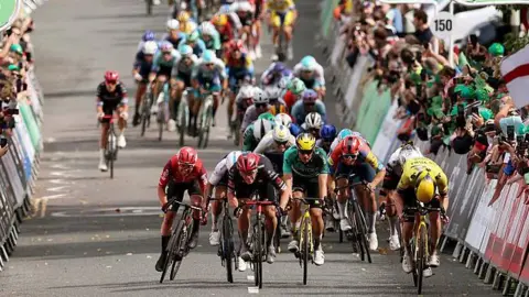 A peloton of cyclists racing on a road, surrounded by crowds of people cheering from behind barriers. 