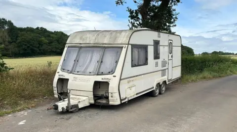 A dirty caravan parked in a country lane with a hedge running alongside and fields in the background