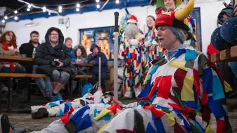 Brook Wassall An actor wearing a white uniform with colourful strips and a horned hat sits on the ground holding a toy sword with onlookers sitting on benches