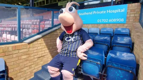 Henry Godfrey-Evans/BBC A mascot with a Southend United jersey on, he appears to be a pink, cartoonish shrimp and is sat on some seats at the football ground