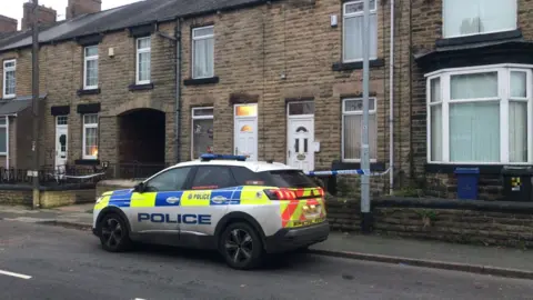 A police car is parked outside a row of terraced houses. One of the houses has blue and white police tape strung outside it. 