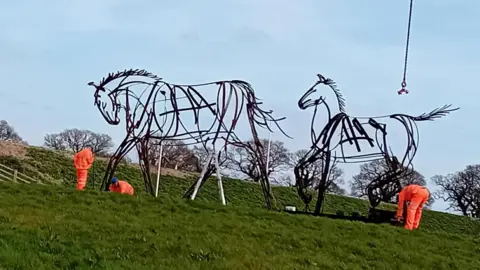 Three workman install two large horst statues. The statues are made of steel and are designed to look as though they have been drawn with pens.