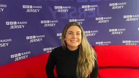 Lesley Mourant sits on a red sofa in front of a Radio Jersey branded purple wall, her long hair is over one shoulder and she smiles towards the camera.