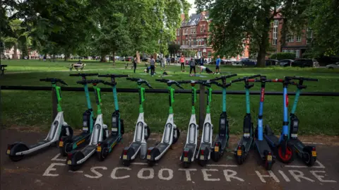 Reuters A row of e-scooters with "E scooter for hire" written in paint on the concrete they are on, with the backdrop of a green space with people doing a yoga session on the grass, surrounded by trees and buildings beyond.