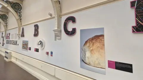 Ruth Millington Interior of a museum or gallery space with arched metal supports and a glass ceiling, featuring a wall display titled “A–Z Birmingham” with large decorative letters, informational panels, and a photograph of a bread roll.
