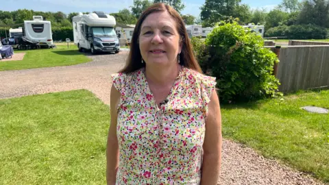 Maureen Bessant standing in front of several camper vans at Greenacres Campsite. She is wearing a pink, green and yellow flowery blouse with black dots.